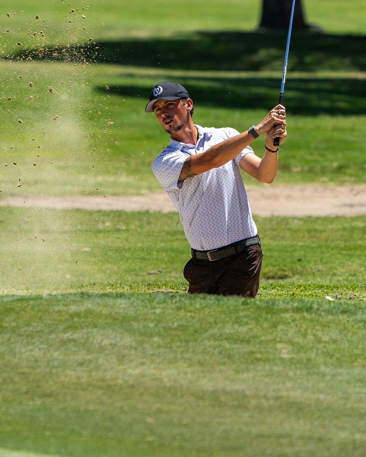 A golfer mid-swing, sand flying, on a bright course